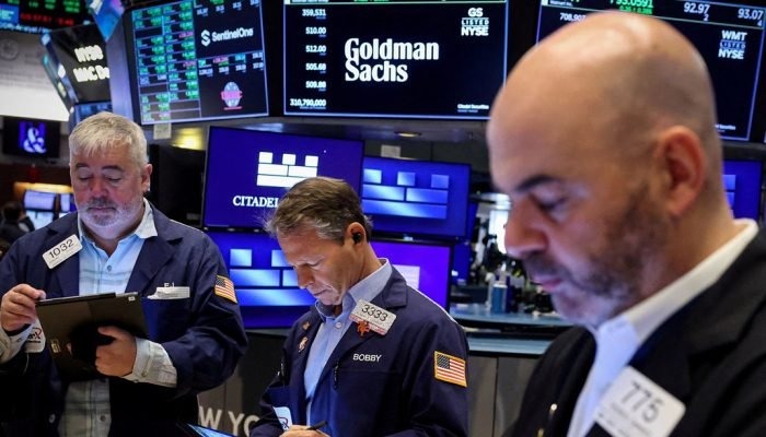 Traders trabalham no pregão enquanto uma tela exibe o logo do Goldman Sachs e informações de n...
