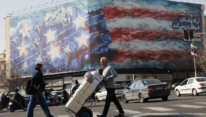 Pessoas caminham em frente a um cartaz anti-EUA em Teerã 19/02/2026 Majid Asgaripour/WANA (West ...